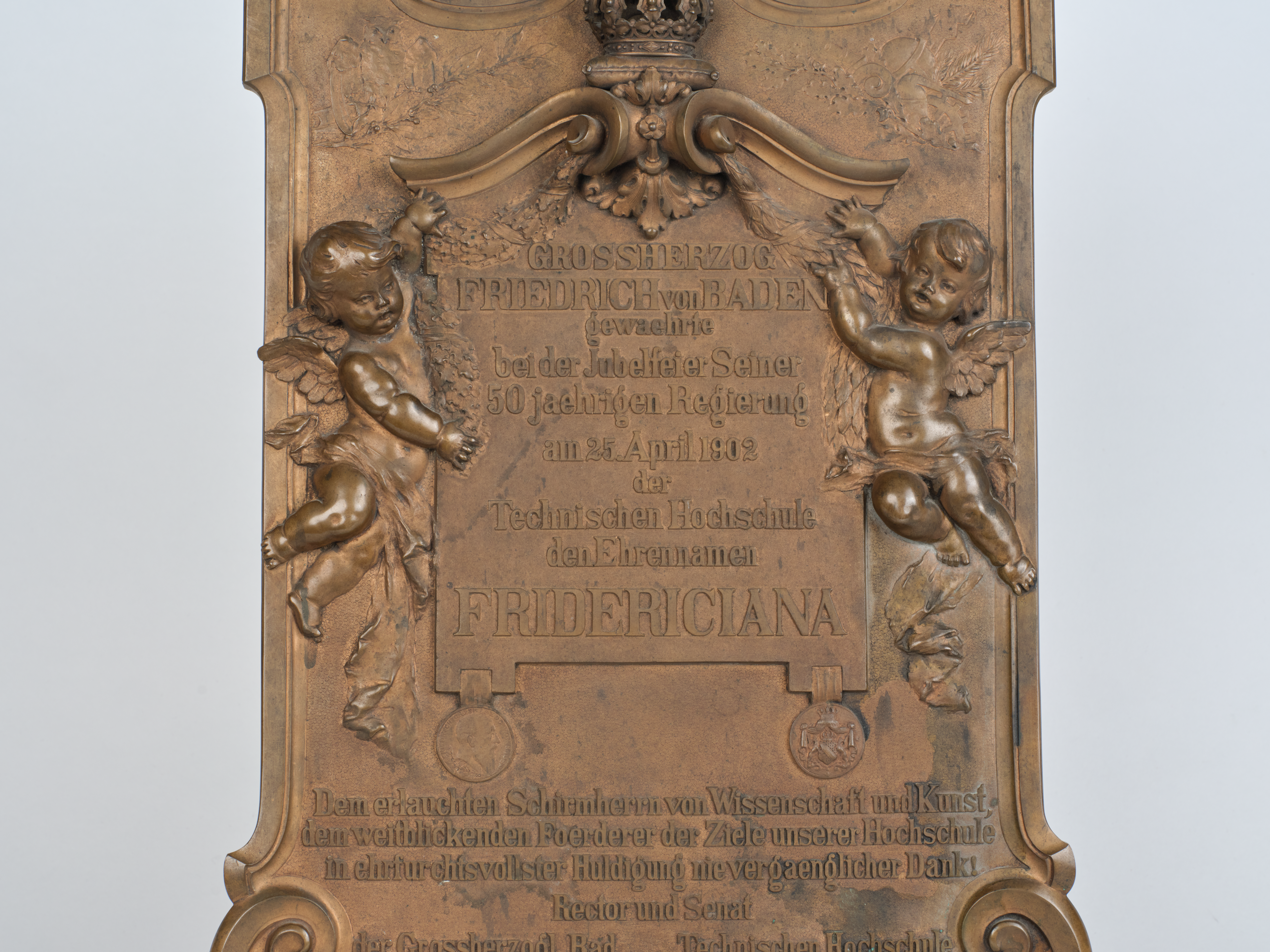 Memorial plaque on the bestowal of the epithet Fridericiana on Karlsruhe Polytechnic, details in the center: plaque held by putti with inscription on the dedication, hanging from it medallions with the portrait of Grand Duke Friedrich I. and the Baden coat of arms. Photograph: Amadeus Bramsiepe and Jonas Zilius.